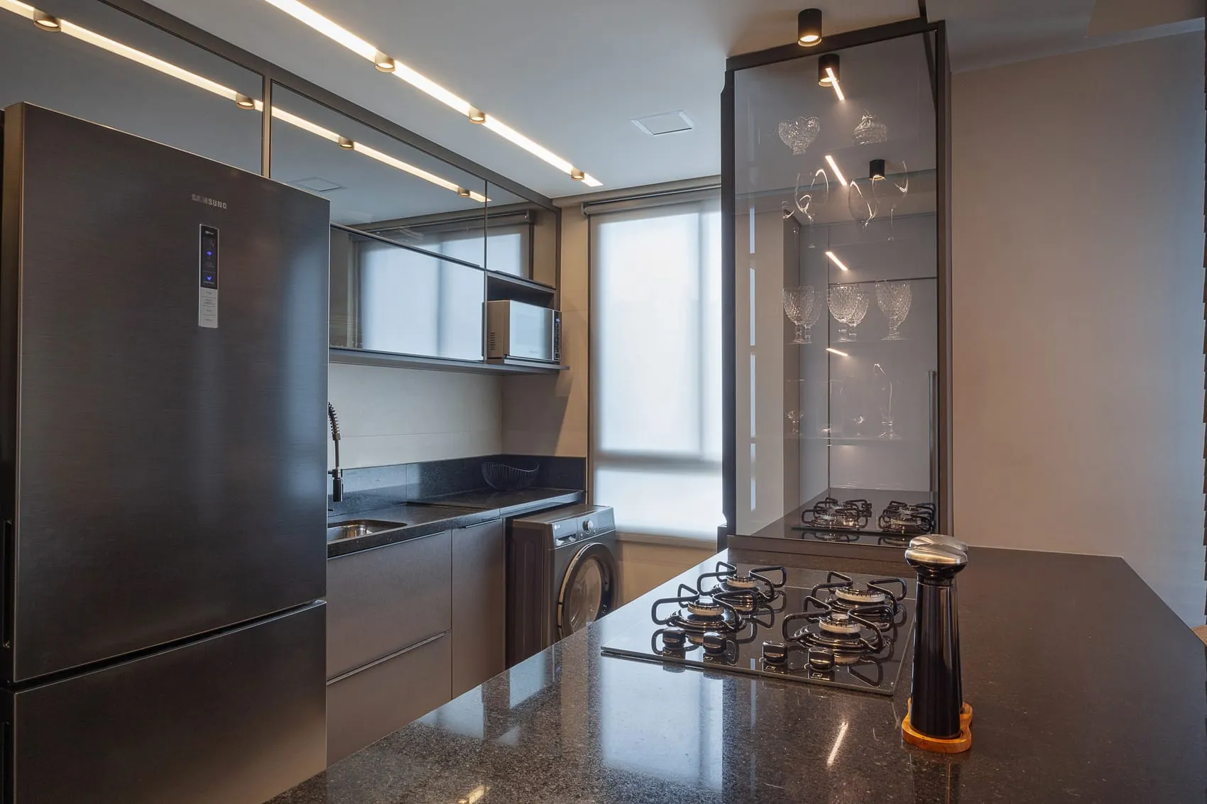 Cozinha com bancada de granito escuro, cooktop e cristaleira de vidro ao fundo