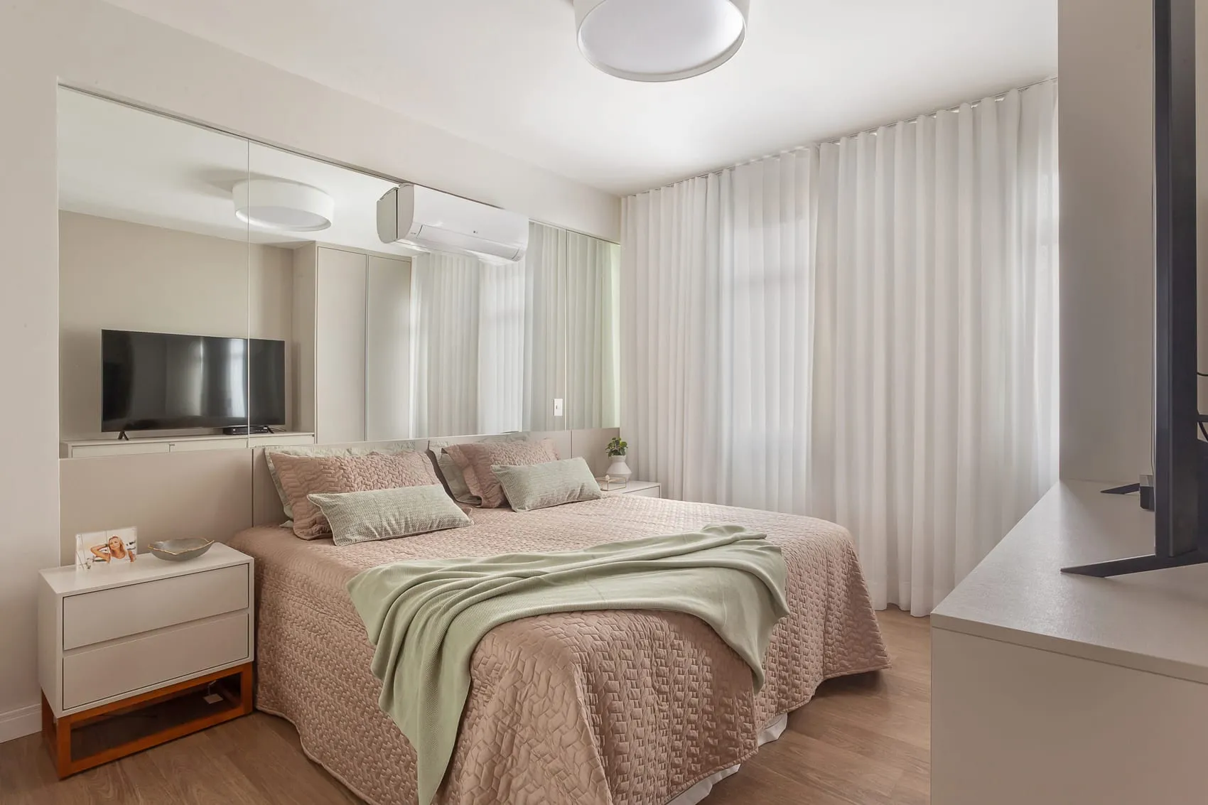 Quarto de casal com roupa de cama verde-clara, cortina e televisão na parede