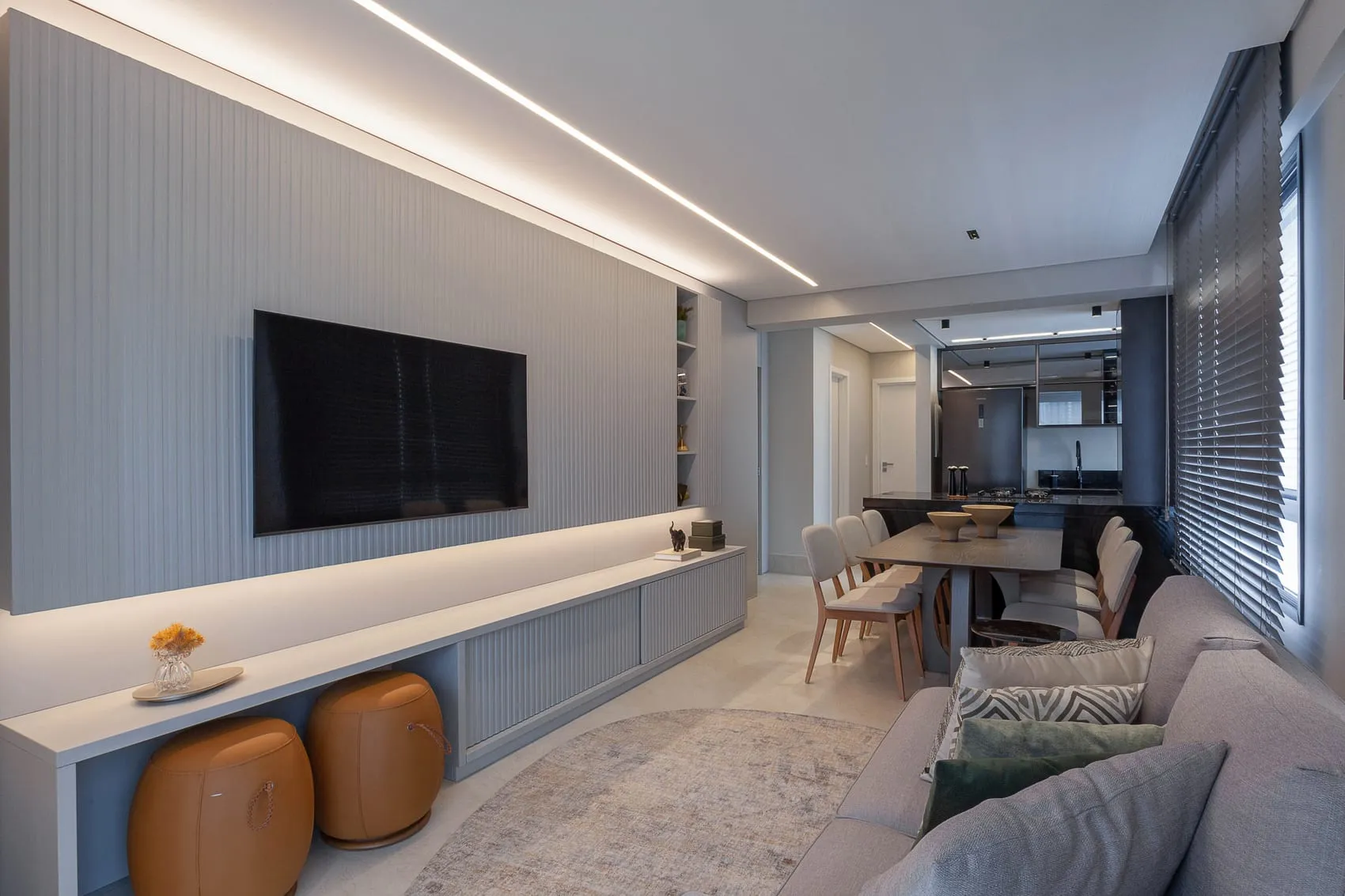 Sala de estar com painel de TV, banquetas de couro e sala de jantar ao fundo