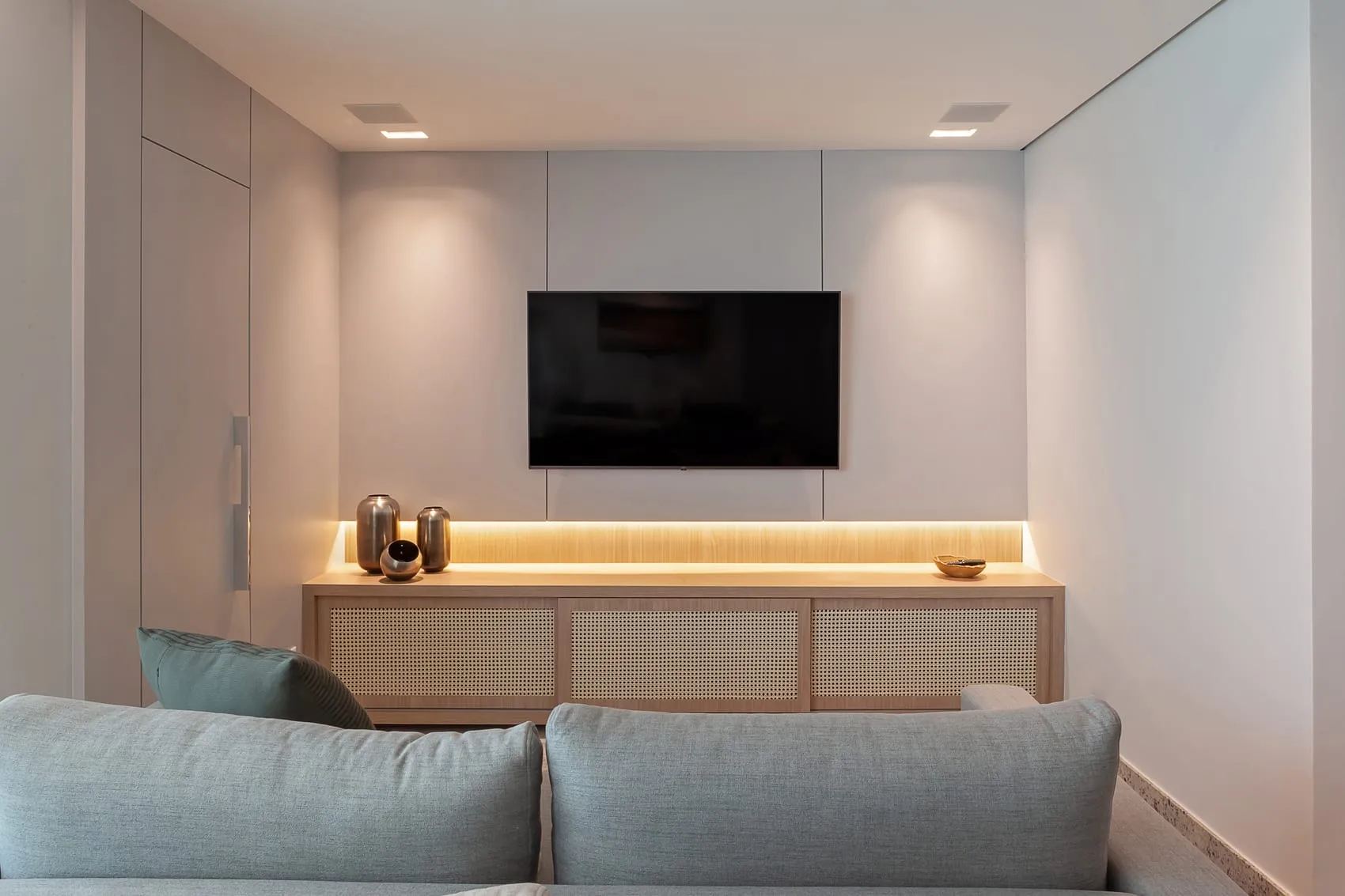 Painel de TV com marcenaria clara, iluminação embutida e sofá azul-acinzentado