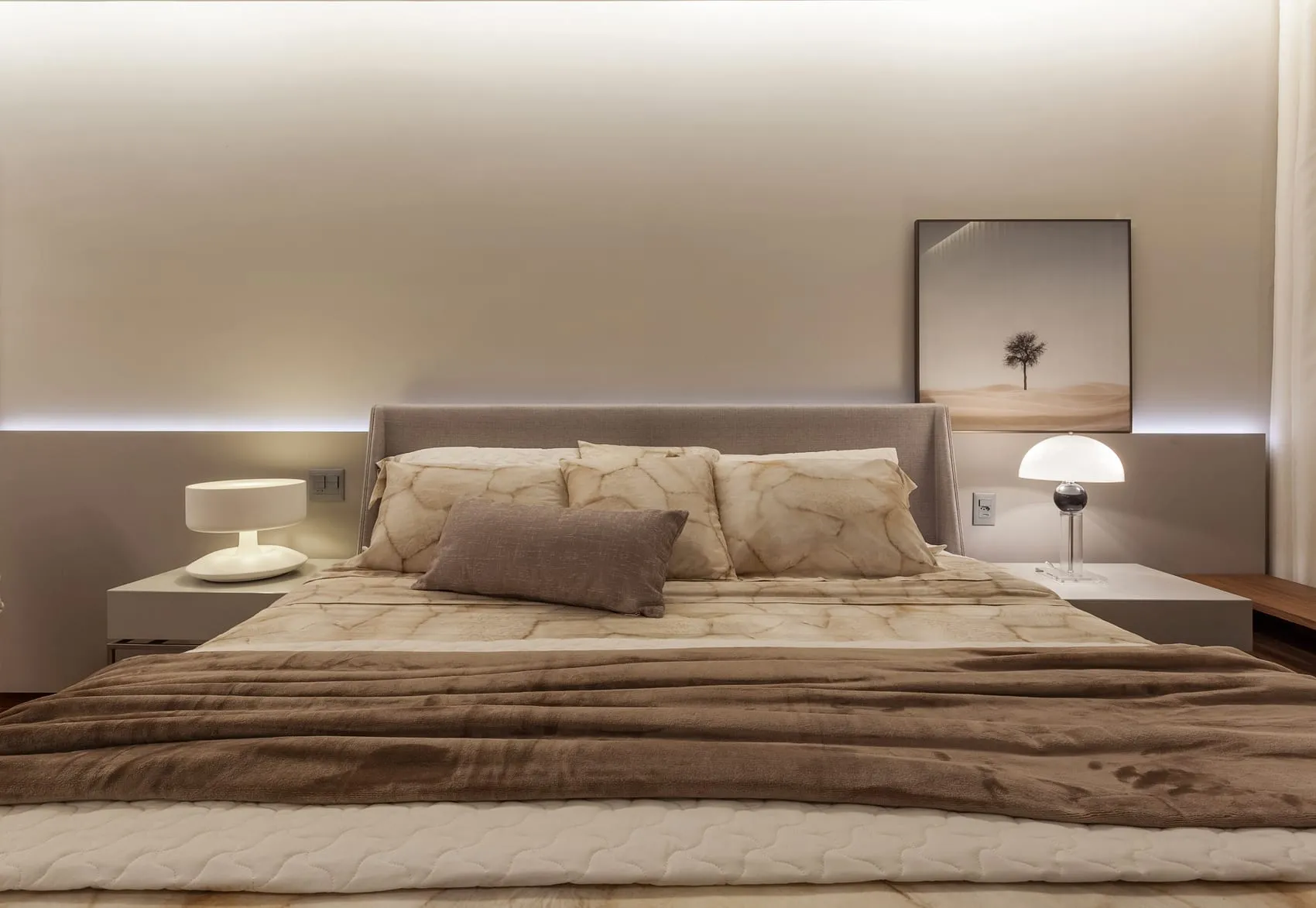 Cama de casal vista de frente, com roupa de cama em tons de marrom e luminárias de mesa