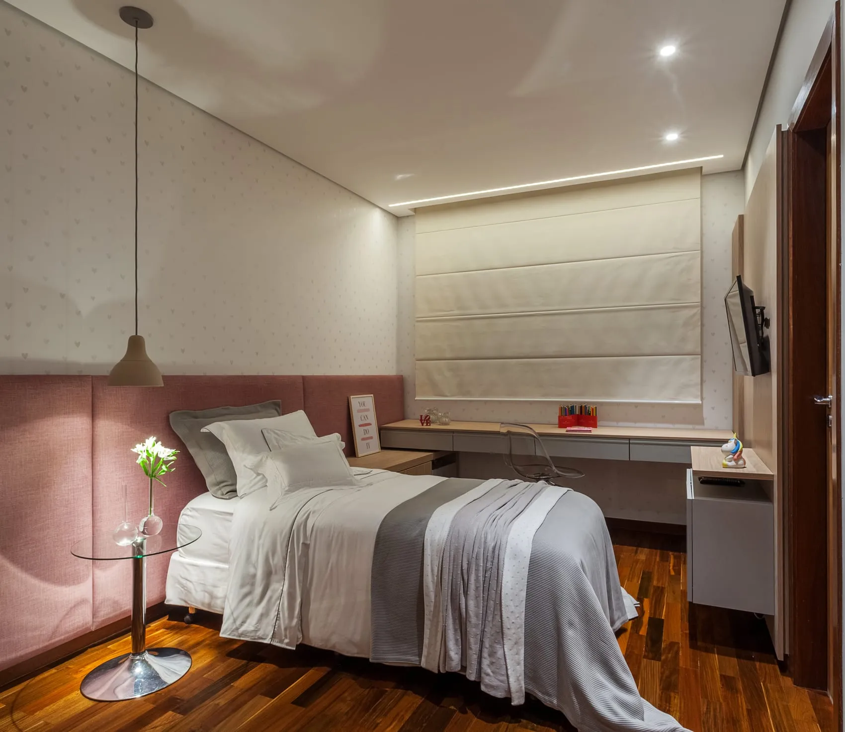 Quarto de solteiro com cabeceira rosa, bancada de estudos e piso de madeira