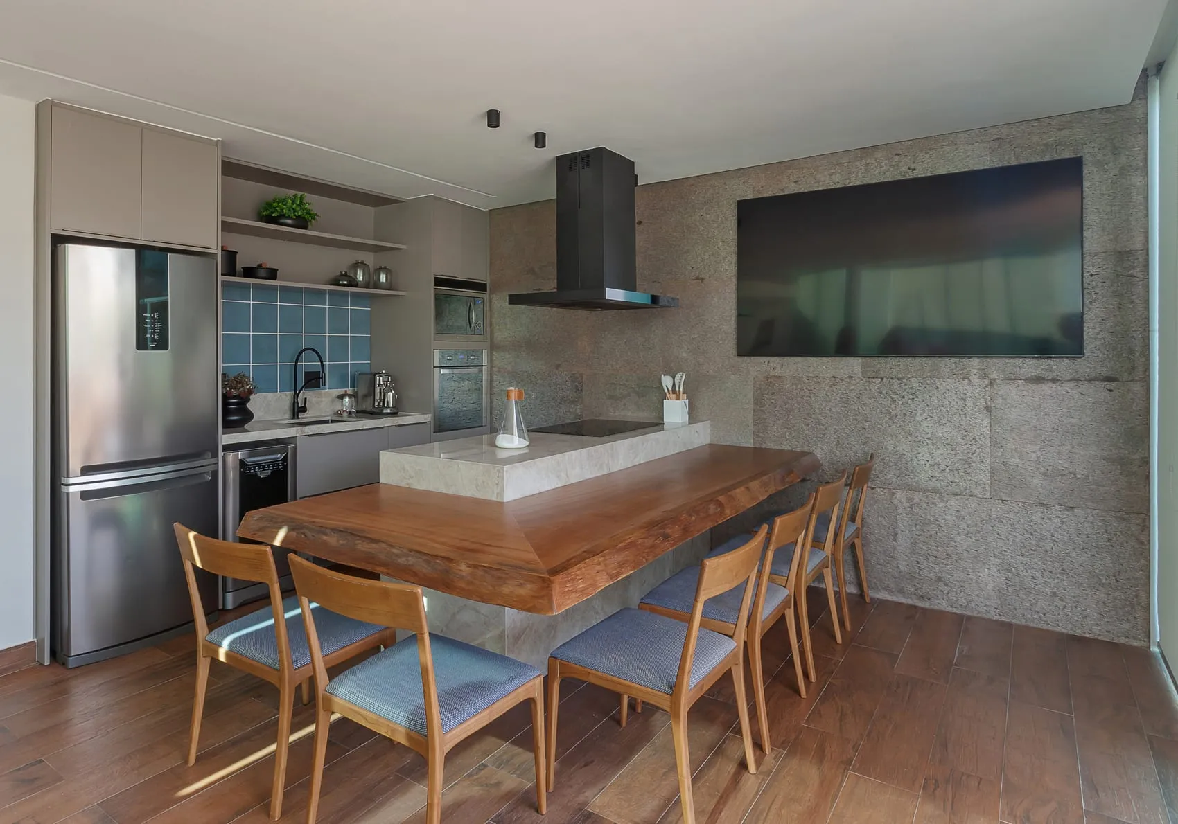 Área de refeições com mesa de madeira, cadeiras azuis e parede de concreto