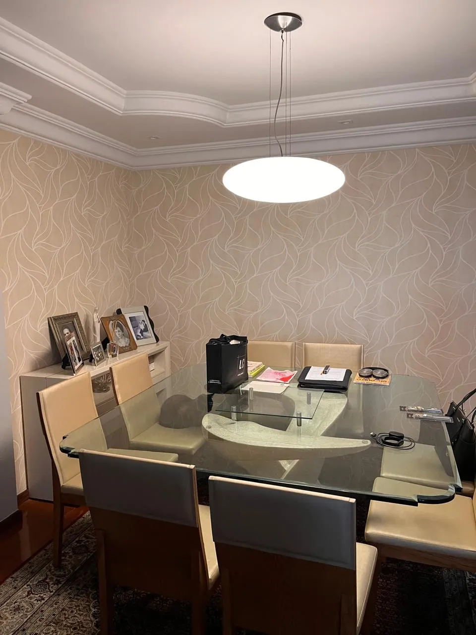 Sala de jantar antiga com mesa de vidro, papel de parede claro e objetos acumulados