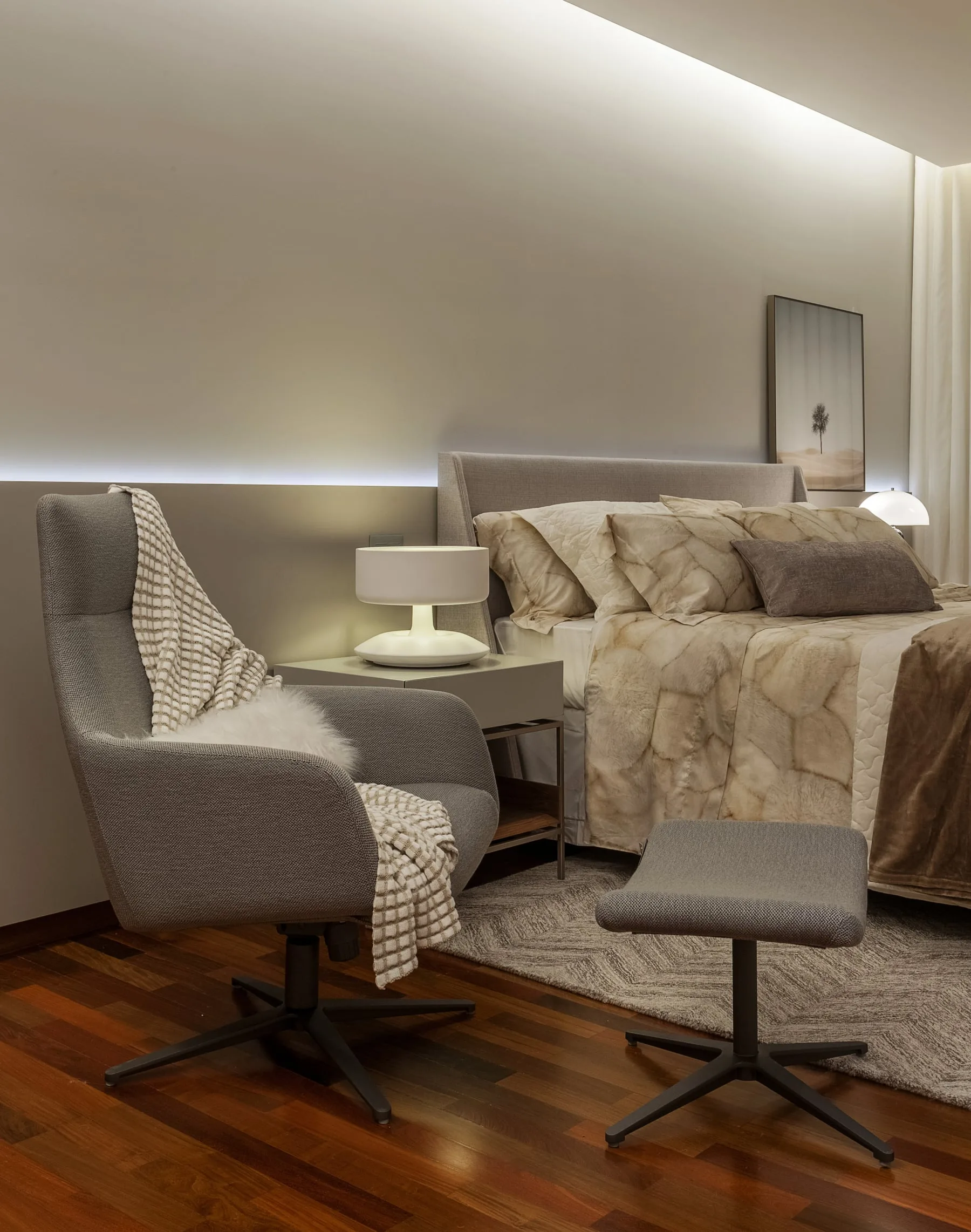 Quarto com cabeceira estofada clara, poltrona, luminárias de mesa e roupa de cama em tons neutros