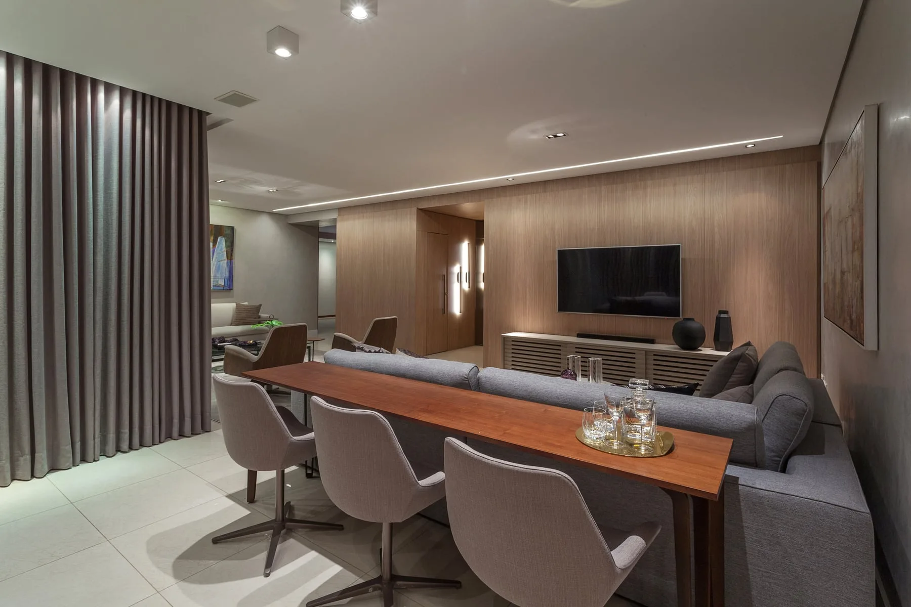 Sala integrada com painel de madeira, mesa de jantar alongada e cortinas claras